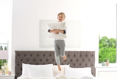 Cute little boy with pillow jumping on bed at home