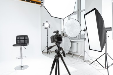 Photo of Tripod with camera, bar stool and professional lighting equipment in modern photo studio