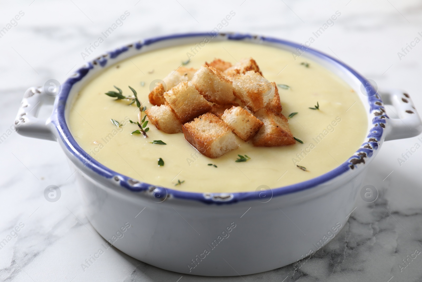 Photo of Tasty potato soup with croutons and rosemary in ceramic pot on white marble table, closeup