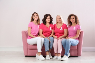 Photo of Group of women with silk ribbons sitting on sofa against light wall. Breast cancer awareness concept