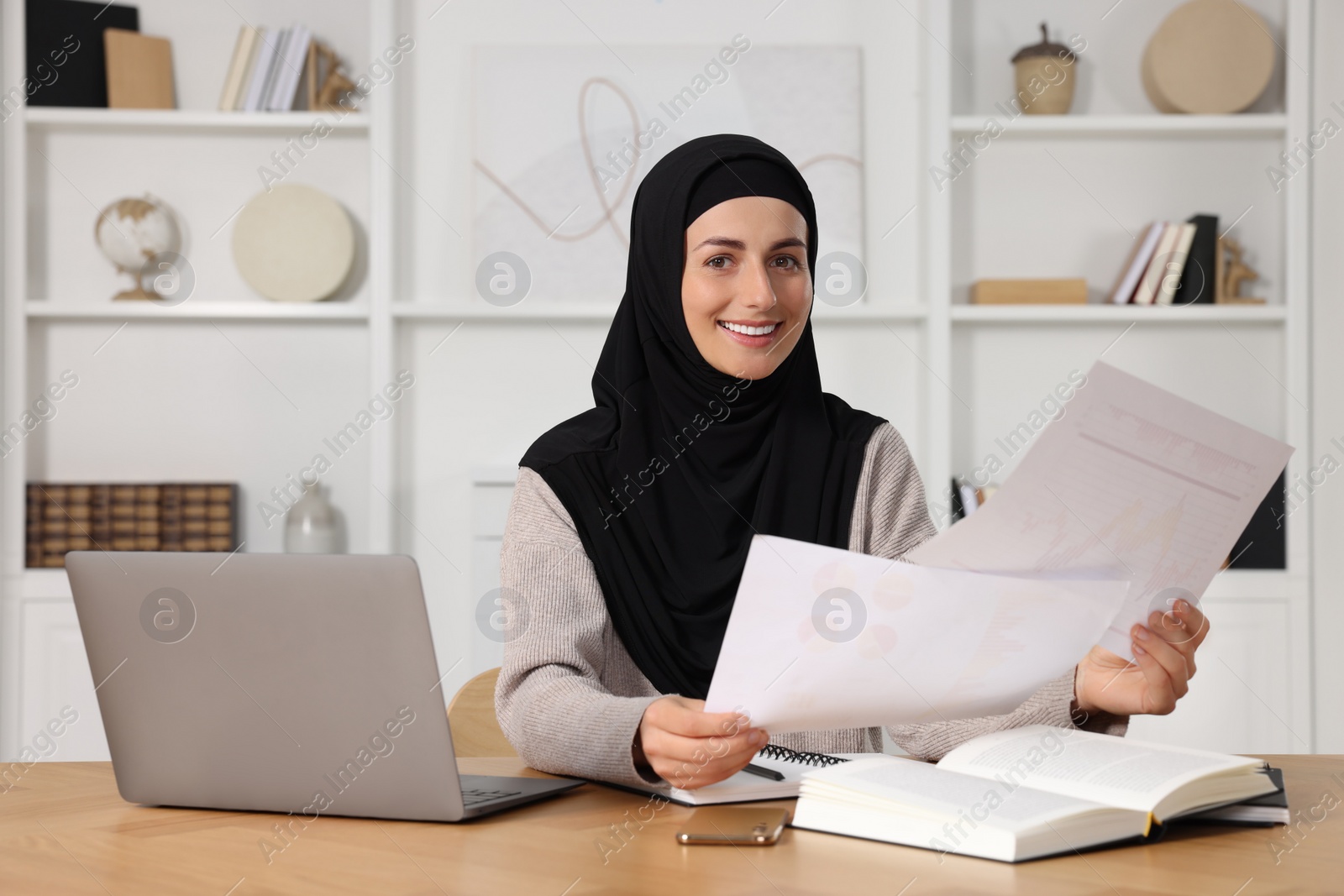 Photo of Muslim woman working near laptop at wooden table in room