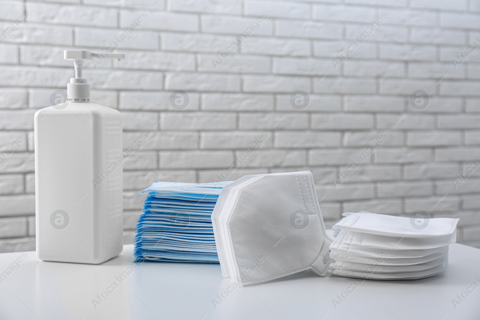 Photo of Hand sanitizer and respiratory masks on table near white brick wall. Protective essentials during COVID-19 pandemic