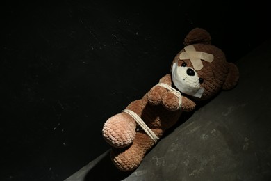 Stop child abuse. Tied toy bear with taped mouth and patches lying on grey floor against black background. Space for text