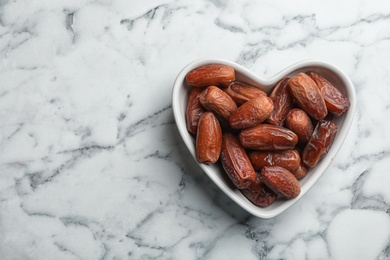 Photo of Heart shaped bowl with sweet dried date fruits on marble background, top view. Space for text