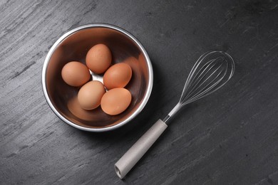 Photo of Metal whisk and raw eggs in bowl on dark grey table, flat lay