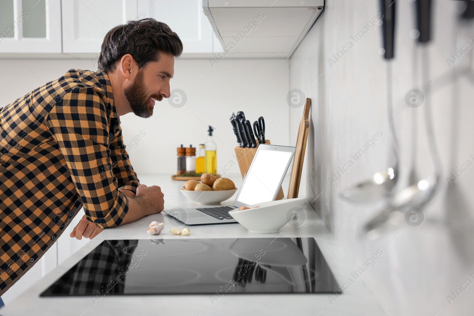 Photo of Man making dinner while watching online cooking course via laptop in kitchen