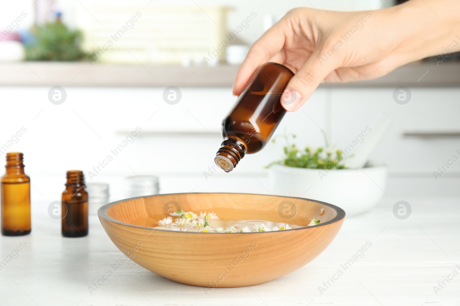 Photo of Woman dripping essential oil into bowl with water and flowers on table