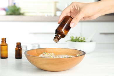 Photo of Woman dripping essential oil into bowl with water and flowers on table