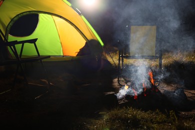 Photo of Smoking bonfire and folding chairs near camping tent outdoors in evening