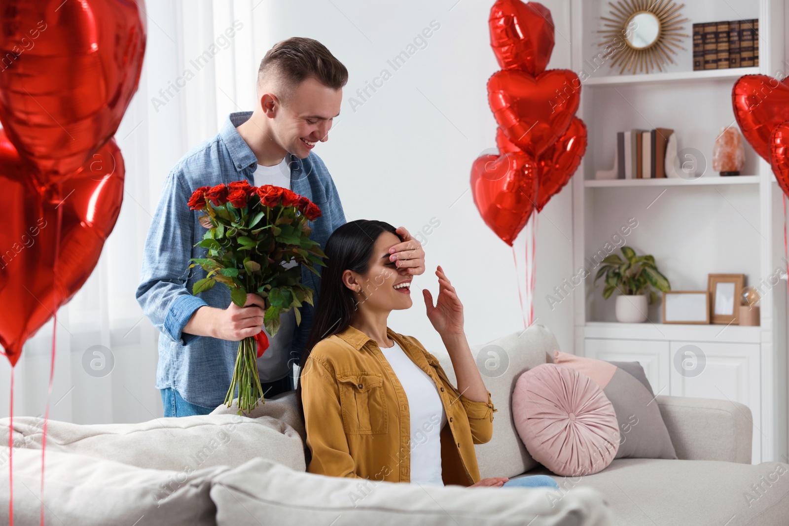 Photo of Boyfriend presenting beautiful bouquet of roses to his girlfriend in room decorated with heart shaped balloons. Valentine's day celebration