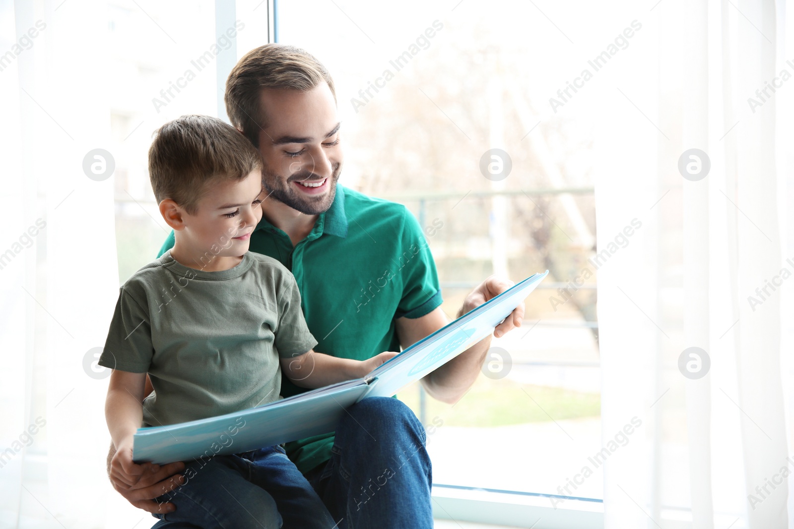 Photo of Young man and his little son reading book near window at home