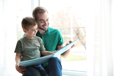 Young man and his little son reading book near window at home