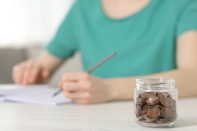 Photo of Financial savings. Woman making notes at white wooden table indoors, focus on glass jar with coins