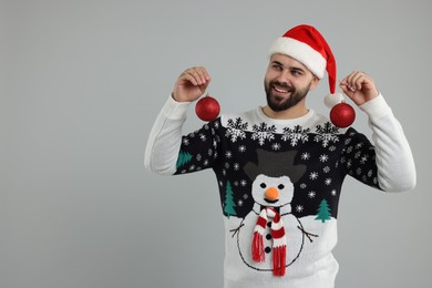 Photo of Happy young man in Christmas sweater and Santa hat holding festive baubles on grey background. Space for text