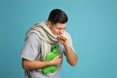 Photo of Ill man with hot water bottle and knitted scarf coughing on light blue background
