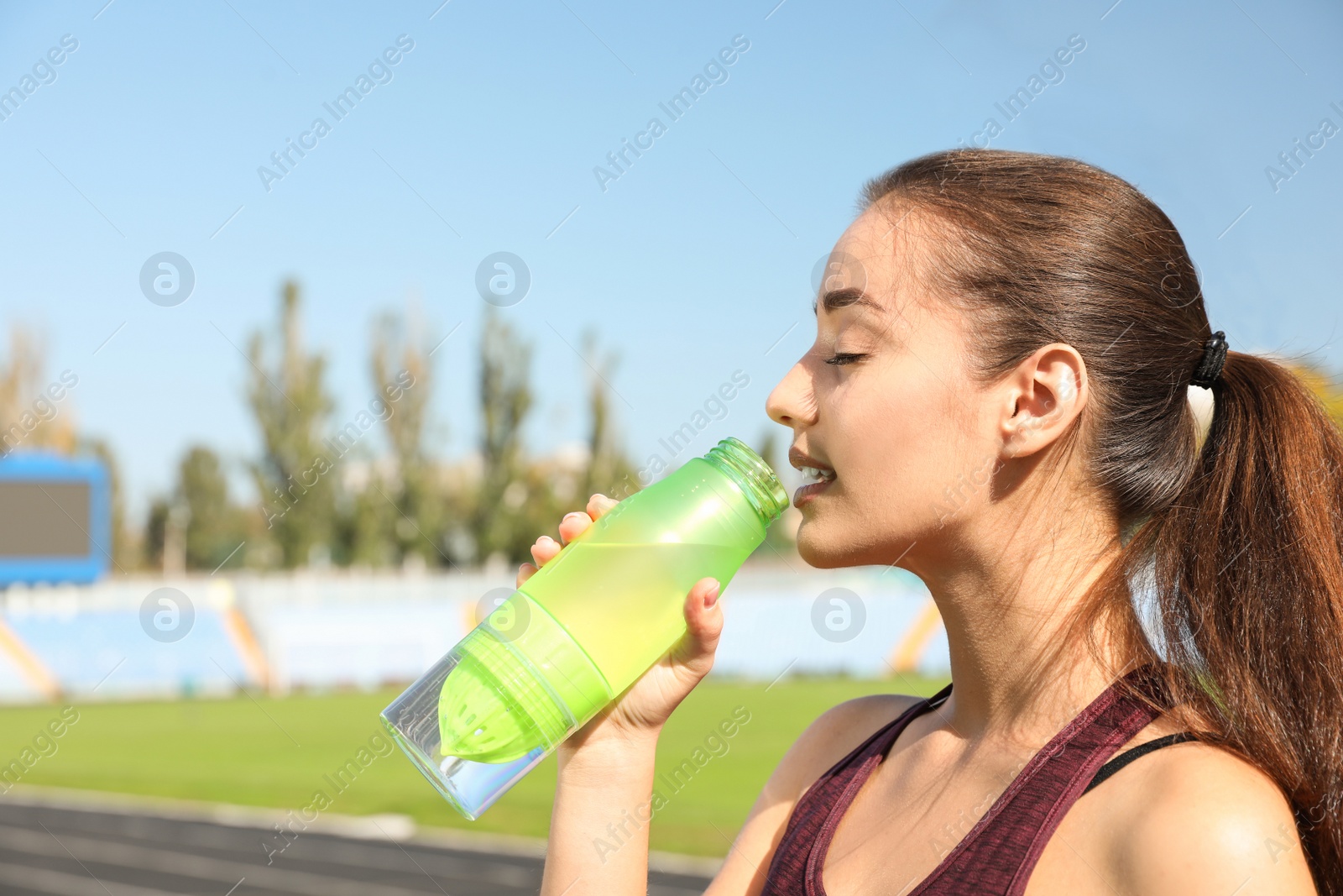 Photo of Young sporty woman drinking water from bottle at stadium on sunny day. Space for text