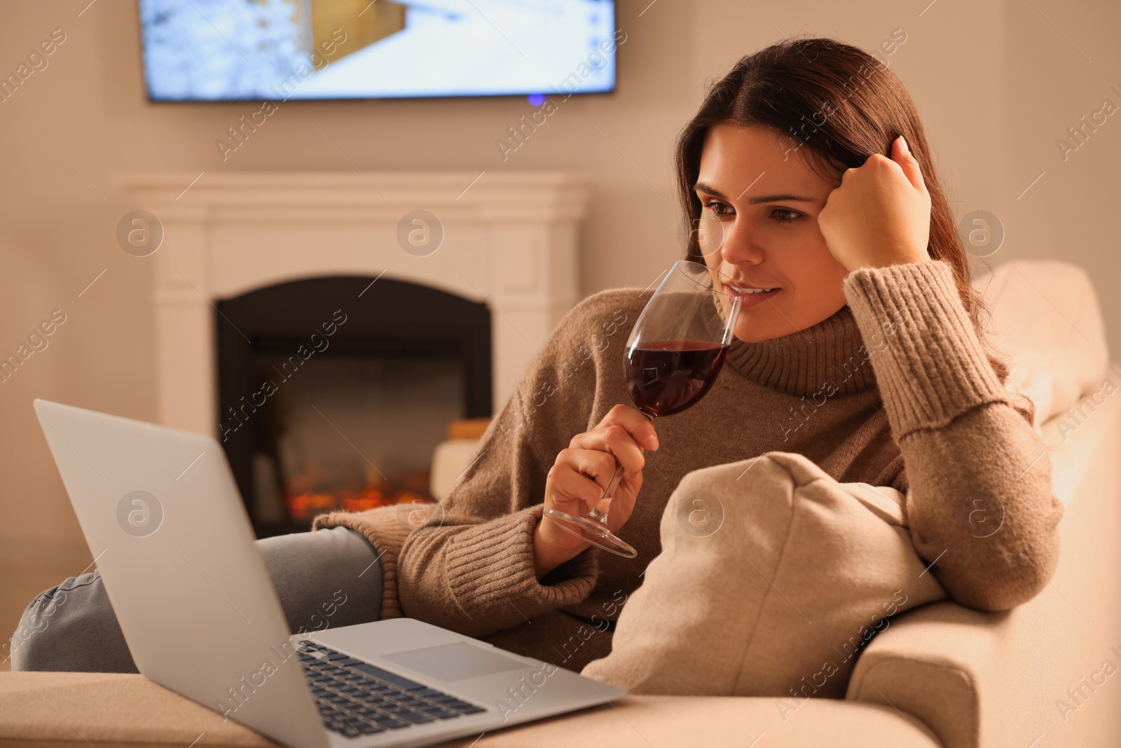 Photo of Young woman with glass of wine and laptop near fireplace at home