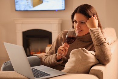 Young woman with glass of wine and laptop near fireplace at home