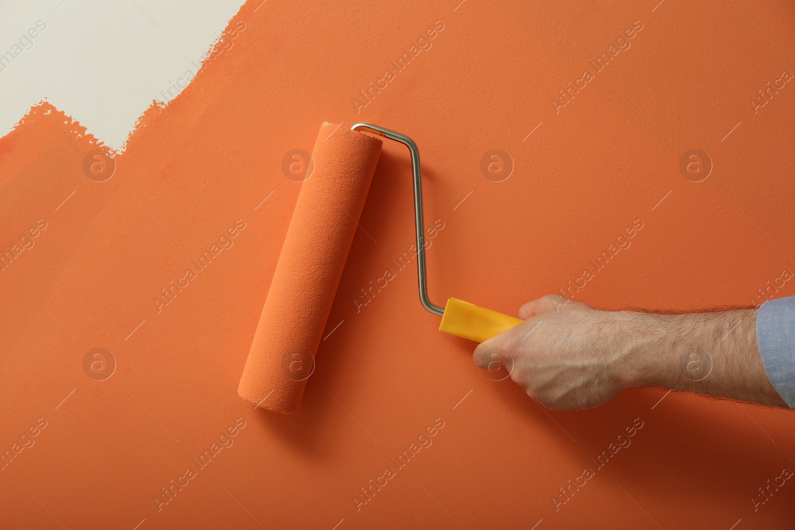 Photo of Man applying orange paint with roller brush on white wall, closeup