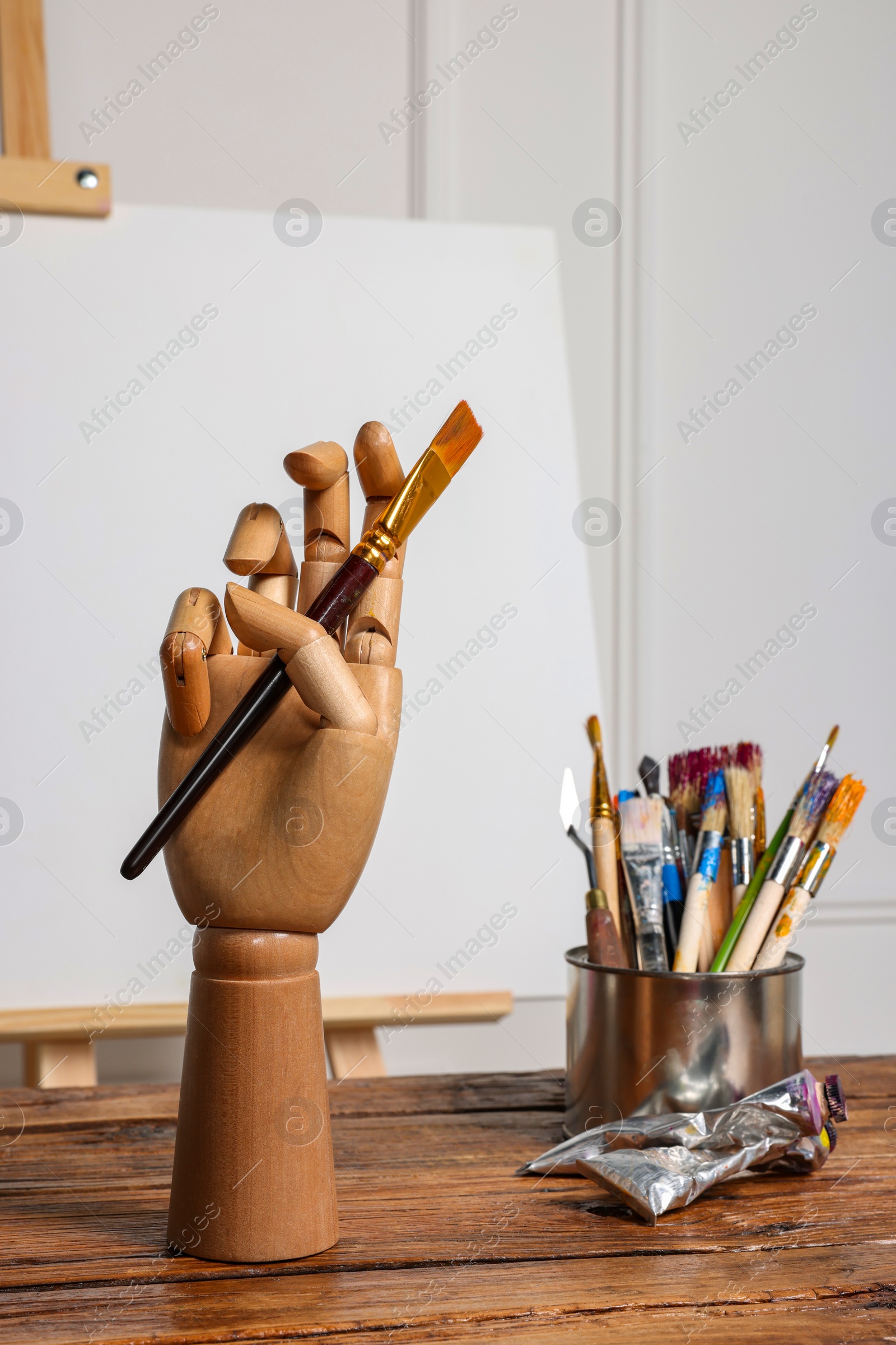 Photo of Easel with blank canvas, hand model and different art supplies on wooden table near white wall