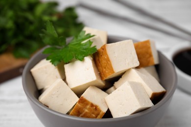 Bowl of smoked tofu cubes, soy sauce and parsley on white wooden table, closeup