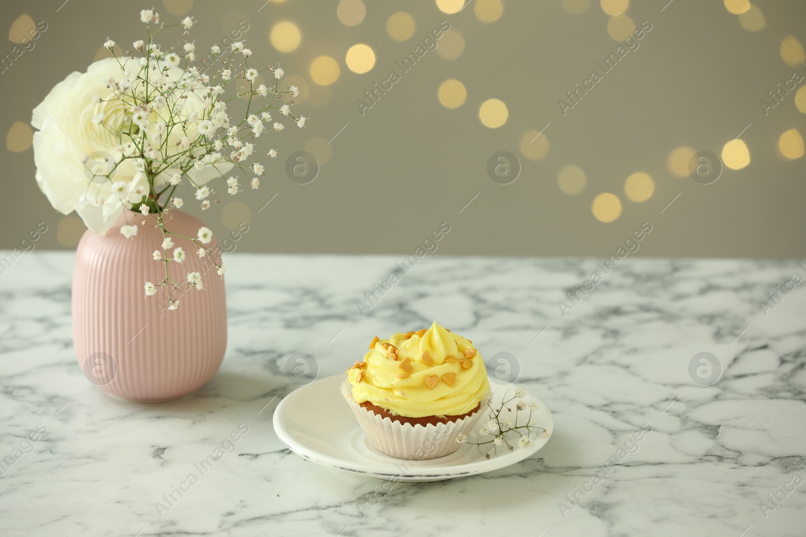 Photo of Delicious cupcake with yellow cream and flowers on white marble table against blurred lights