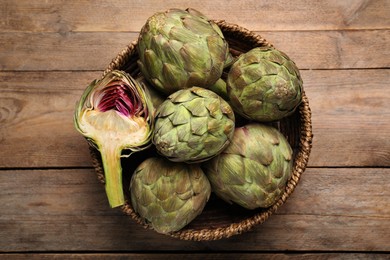 Photo of Cut and whole fresh raw artichokes in basket on wooden table, top view