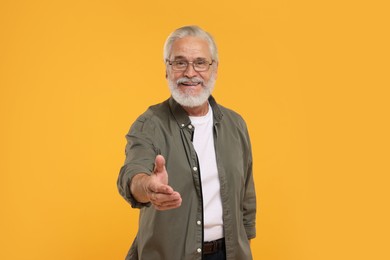 Photo of Senior man in glasses welcoming and offering handshake on yellow background