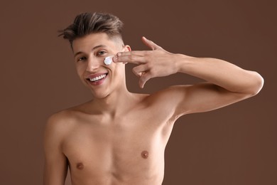 Handsome man applying moisturizing cream onto his face on brown background