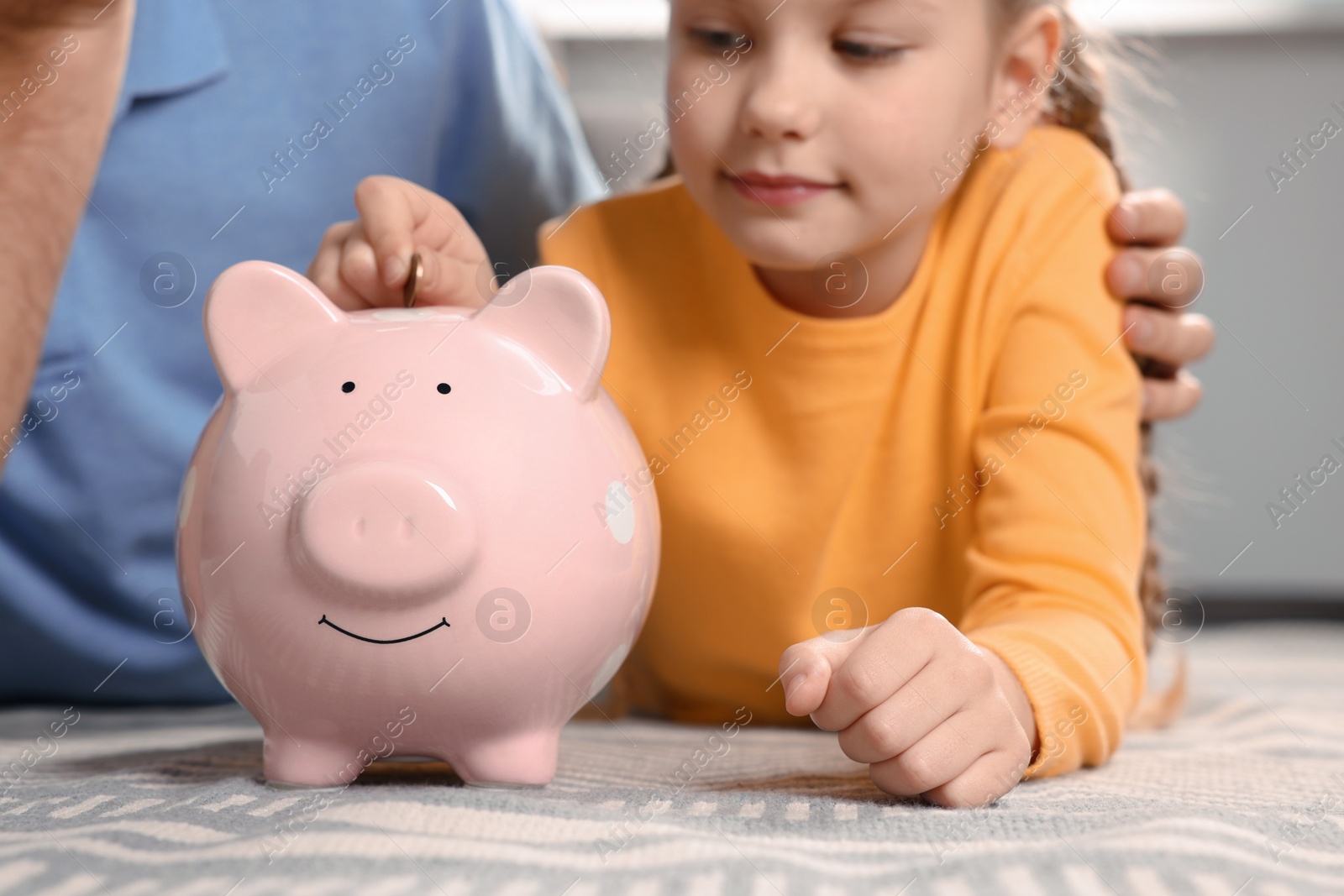 Photo of Little girl with her father putting coin into piggy bank at home, closeup