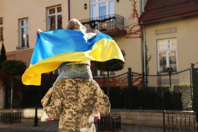 Soldier in military uniform carrying his daughter on shoulders and Ukrainian flag on city street, back view with space for text. Family reunion