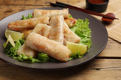 Photo of Plate with tasty fried spring rolls, lettuce, lime and chopsticks on wooden table, closeup