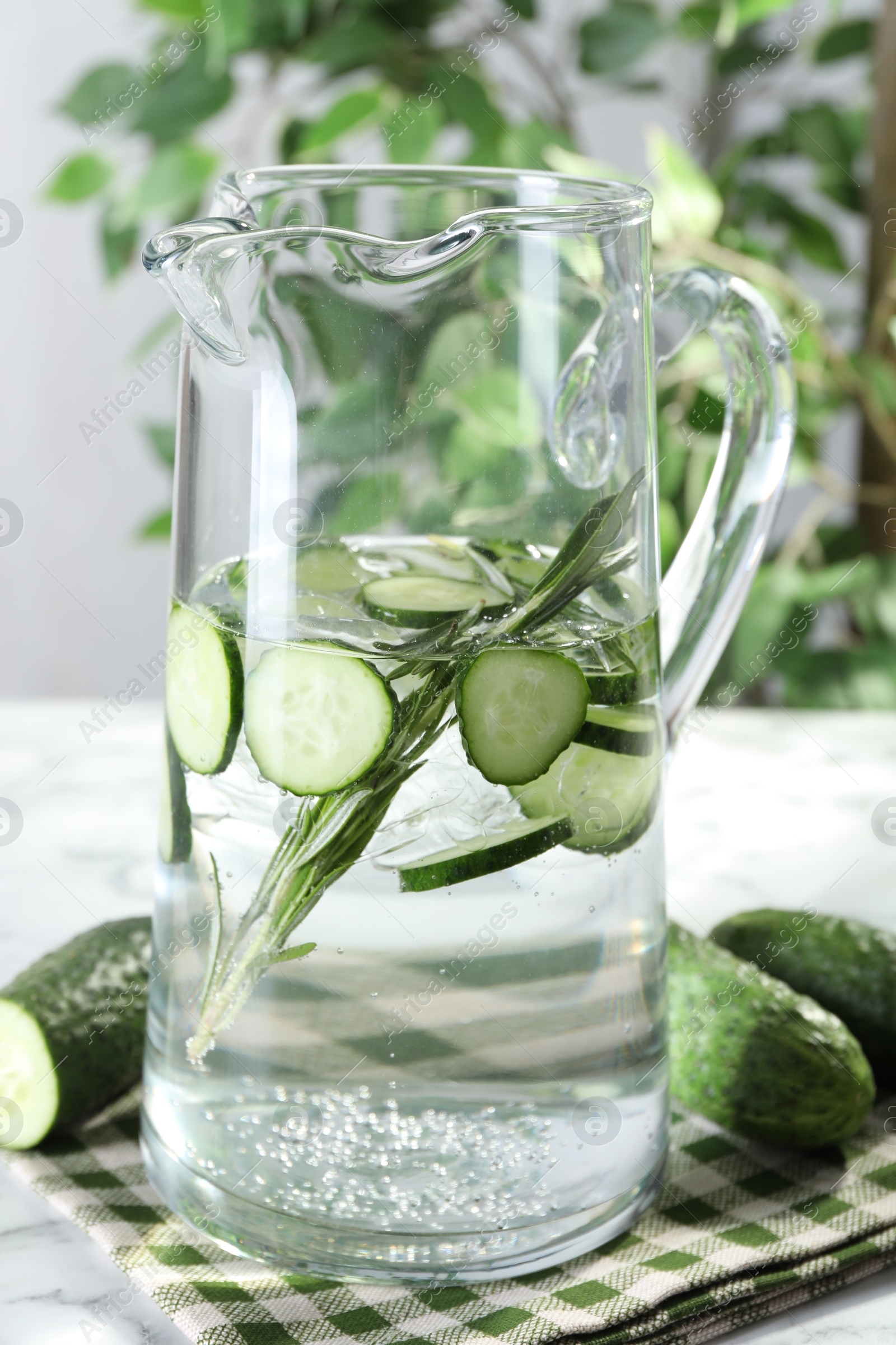 Photo of Refreshing cucumber water with rosemary in jug and vegetables on table, closeup