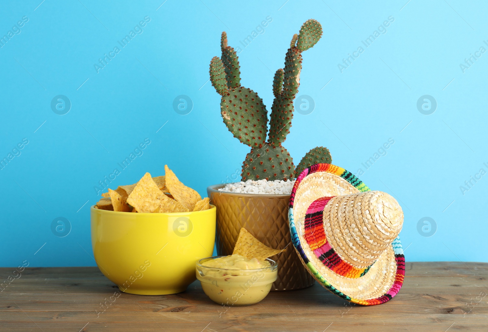 Photo of Mexican sombrero hat, cactus, nachos chips and guacamole in bowls on wooden table against light blue background