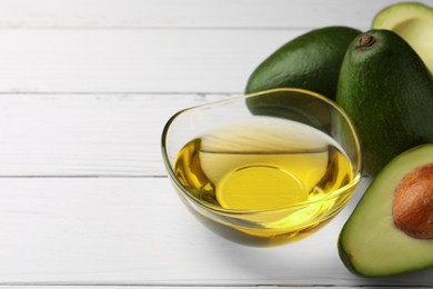 Cooking oil in bowl and fresh avocados on white wooden table, space for text