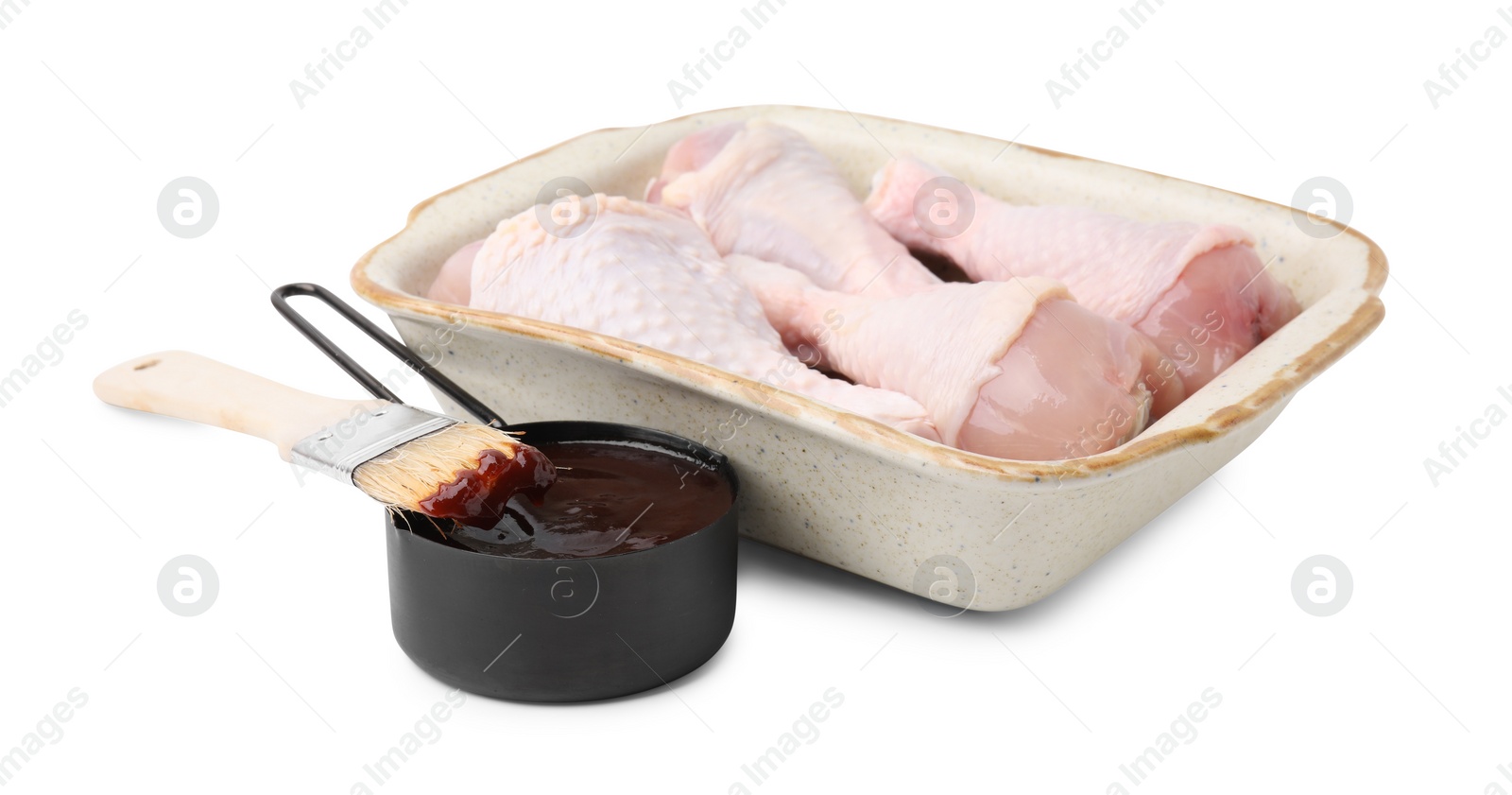 Photo of Raw chicken drumsticks in baking dish, marinade and basting brush isolated on white