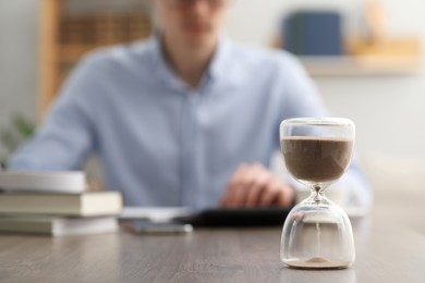 Photo of Hourglass with flowing sand on desk. Man using calculator indoors, selective focus