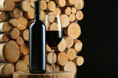 Photo of Stylish presentation of red wine in bottle and wineglass near wooden logs on black background, space for text