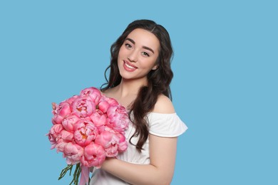 Beautiful young woman with bouquet of pink peonies on light blue background