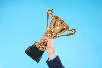 Photo of Businessman holding gold trophy cup on light blue background, closeup