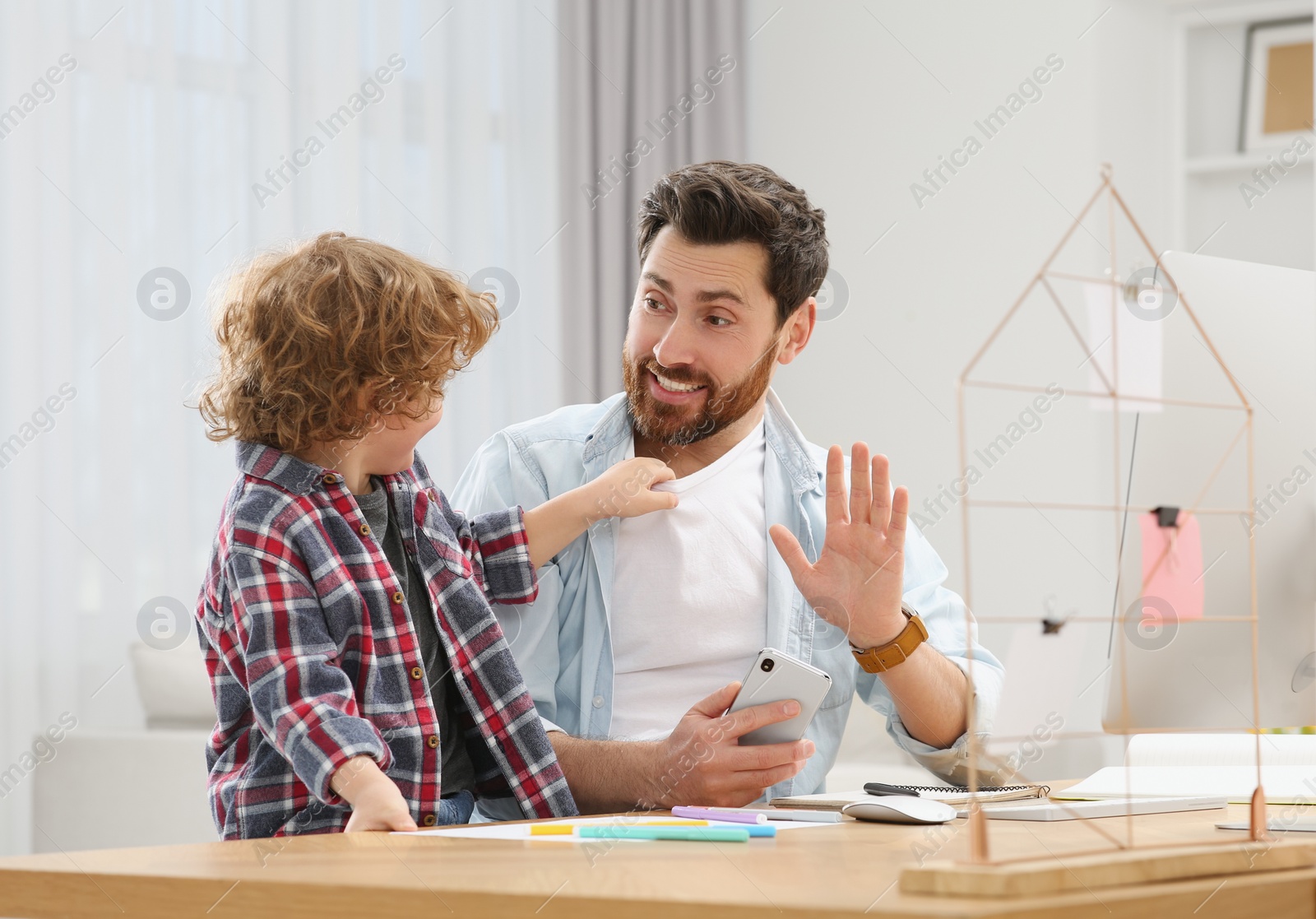 Photo of Little boy bothering his father at home. Man working remotely at desk