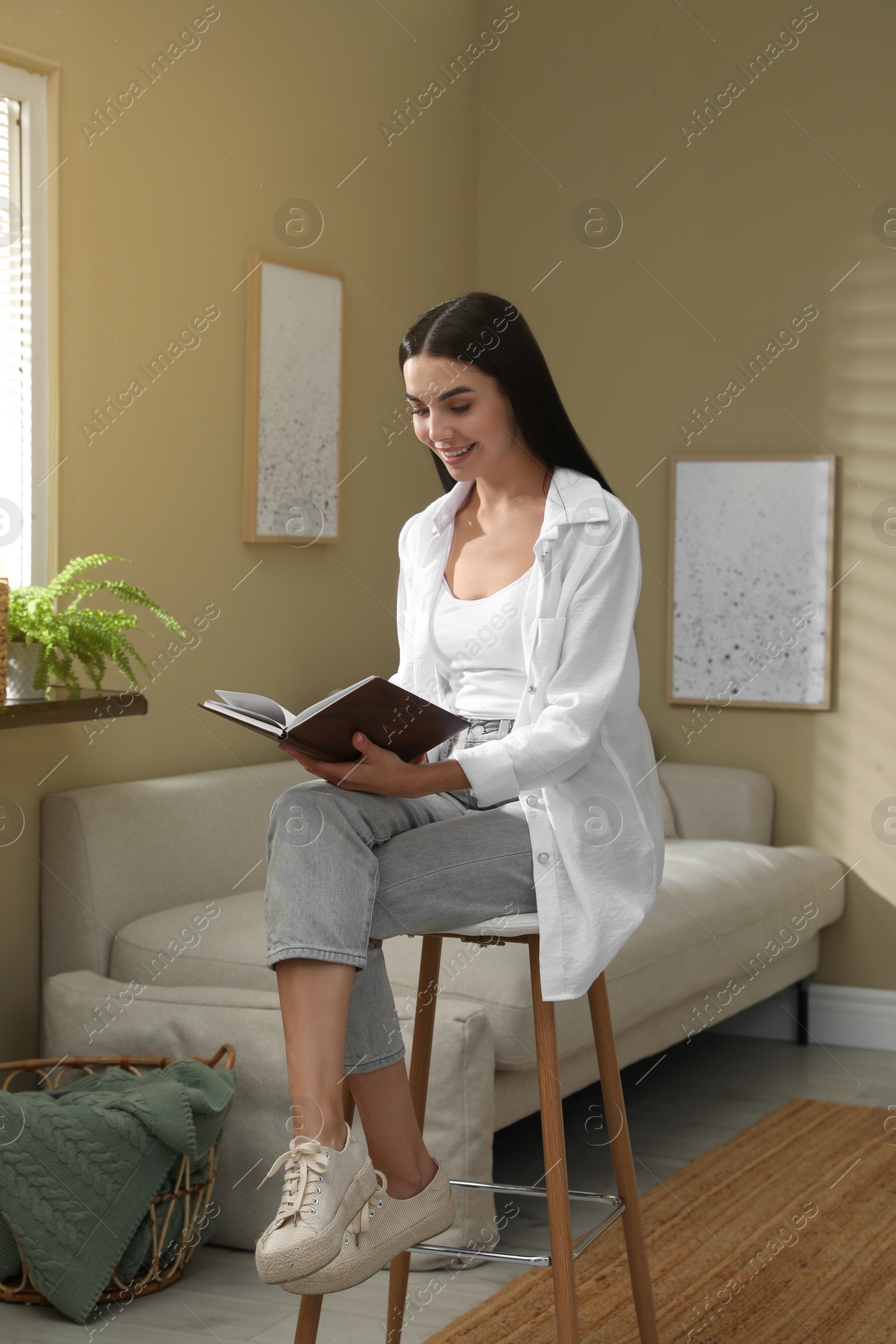 Photo of Beautiful young woman reading book on stool at home