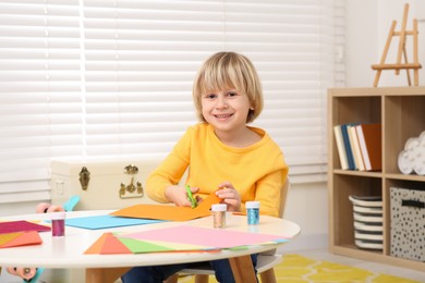 Photo of Cute little boy cutting colorful paper at desk in room. Home workplace