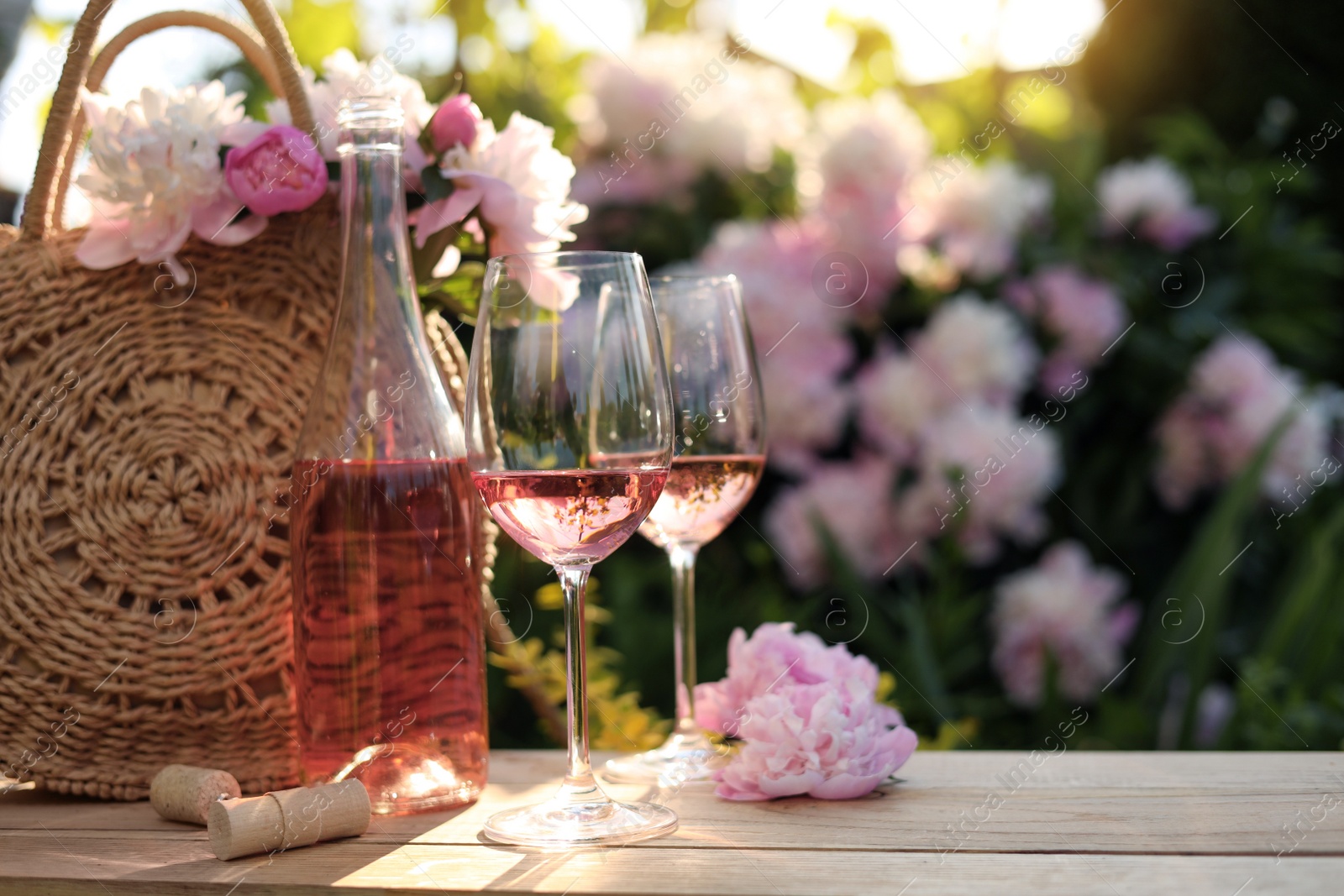 Photo of Bottle and glasses of rose wine near beautiful peonies on wooden table in garden