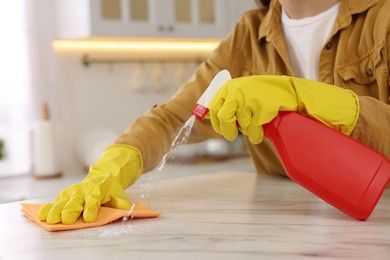 Photo of Woman with spray bottle and microfiber cloth cleaning white marble table in kitchen, closeup