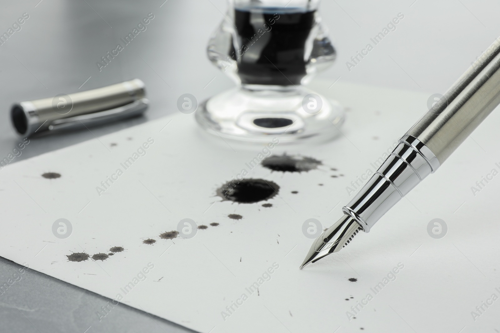 Photo of Stylish fountain pen, paper with blots of ink and inkwell on light grey table