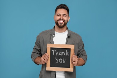 Photo of Happy man holding small chalkboard with phrase Thank You on light blue background