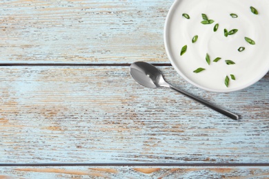 Bowl of fresh sour cream with green onion and spoon on light blue wooden table, flat lay. Space for text