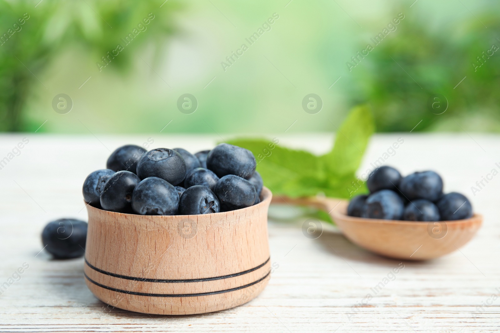 Photo of Wooden bowl and spoon of tasty blueberries on white table against blurred green background, space for text
