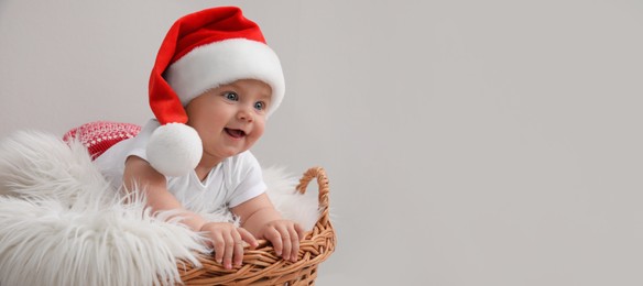 Cute baby wearing Santa hat in wicker basket on light grey background, banner design with space for text. Christmas celebration
